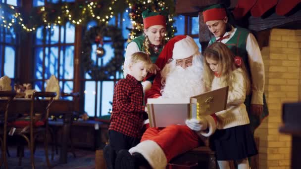 Santa Claus is sitting on a chair surrounded by two elves and a little boy with a girl and considering a holiday album with photos on the background of New Year's decorations and the Christmas tree in the room at home - Πλάνα, βίντεο