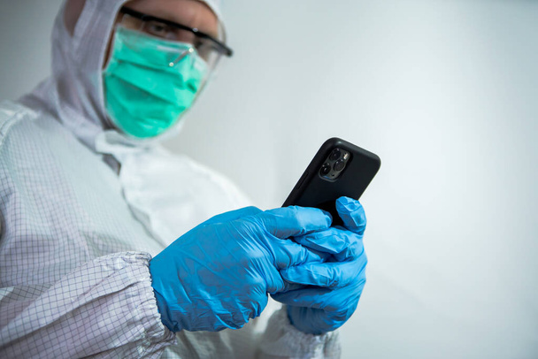 Běloch v ochranném obleku a obličejové masce používající smartphone v laboratoři. Doktor používající telefonní zařízení. Koronavirus způsobil nouzovou situaci. Koncepce pandemické karantény Covid-19 - Fotografie, Obrázek