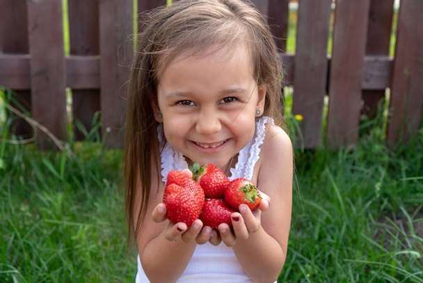 Małe piękne dziecko rozciąga truskawki w dłoniach i uśmiecha się. Urocza biała dziewczyna z długimi czarnymi włosami, trzymająca soczyste truskawki. Koncepcja czasu letniego, dzieciństwa i zdrowia - Zdjęcie, obraz