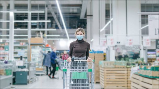 Uma garota séria em uma máscara médica com um carrinho de supermercado está de pé em um supermercado, há multidões de clientes andando por aí. Protecção contra o coronavírus
. - Foto, Imagem