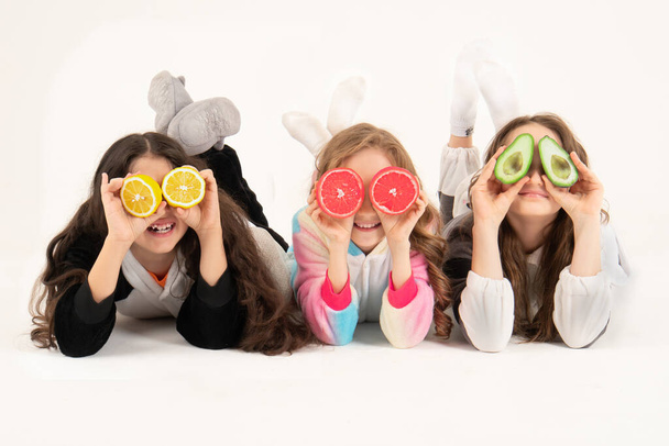 Τρία κορίτσια χρησιμοποιούν φρούτα ως γυαλιά σε λευκό φόντο. Ομάδα χαρούμενων παιδιών με γκρέιπφρουτ, λεμόνι, αβοκάντο στα χέρια - Φωτογραφία, εικόνα