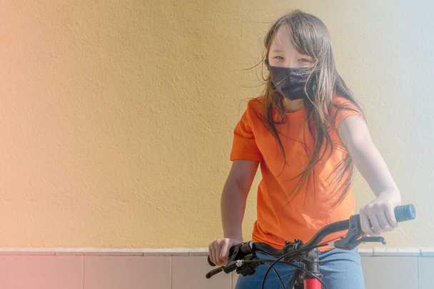 Siyah koruyucu tıbbi maskeli genç kız evin arka planında bisiklete biniyor. Coronavirus ve avluda yürüyor, kendini izole ediyor. - Fotoğraf, Görsel