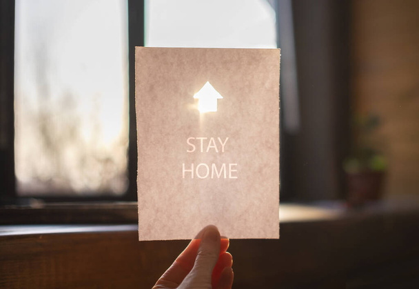 Hoja blanca de papel con un letrero de casa brillante y palabras Quédese en casa en una mano
 - Foto, imagen