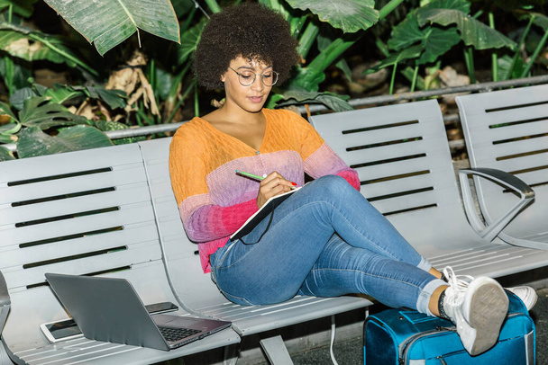 Hermosa mujer de raza mixta escribe en su diario mientras espera con su equipaje sentado en un banco en una estación
 - Foto, imagen
