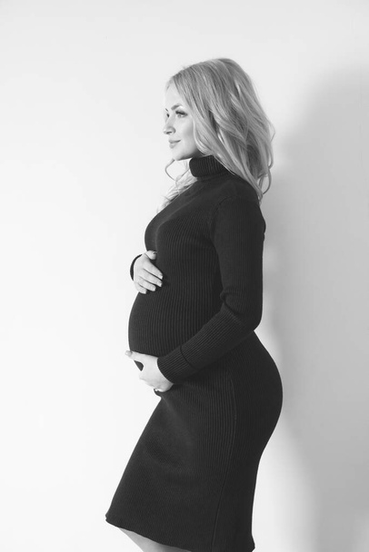Lovely young pregnant woman with blond long hair in a dark dress in the studio. Fashion for pregnant women. Happy motherhood - Foto, afbeelding