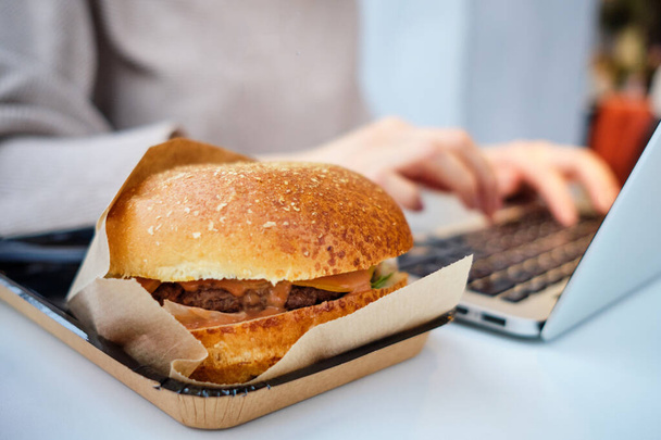 Chica trabajando en un portátil en un restaurante con una hamburguesa. De cerca.
. - Foto, imagen