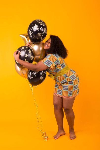 Holidays, birthday party and fun concept - Portrait of smiling young African-American young woman looking sweet on yellow background holding balloons. - Fotoğraf, Görsel
