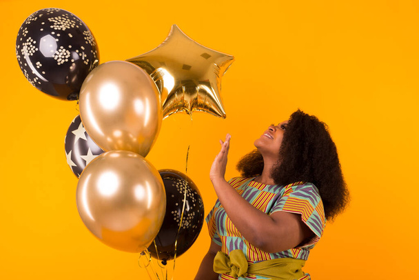Vacances, fête d'anniversaire et concept amusant - Portrait de jeune femme afro-américaine souriante regardant doux sur fond jaune tenant des ballons
. - Photo, image