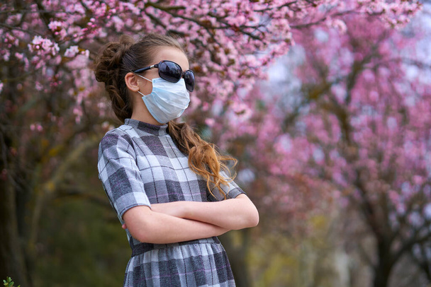 Mädchen mit Gesichtsmaske ist in der Stadt draußen, blühende Bäume, Frühlingszeit, Blütezeit - Konzept für Allergien und Gesundheitsschutz vor staubiger Luft - Foto, Bild