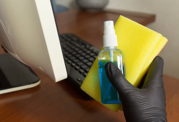 anti-séptico azul em uma garrafa e uma esponja amarela em uma mão em uma luva nitrílica preta. local de trabalho com teclado e computador antes da limpeza
 - Foto, Imagem
