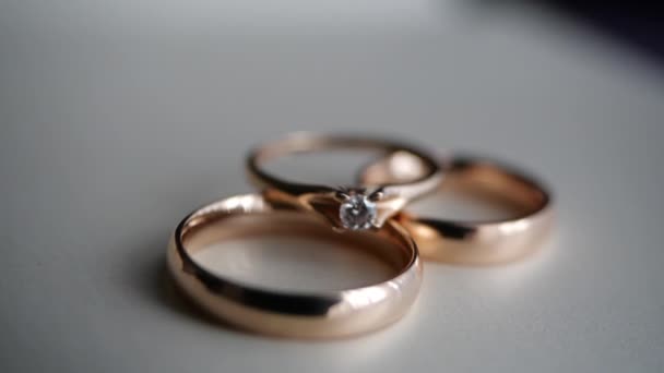 Τρία πανέμορφα χρυσά δαχτυλίδια - ένας αρραβώνας με ένα διαμάντι και μια βέρα κείτονται σε ένα λευκό τραπέζι. Δώρα και ρομαντισμό, διακόσμηση. Εορταστική ατμόσφαιρα της νύφης και του γαμπρού. - Πλάνα, βίντεο