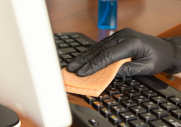hand in a black nitrile glove wipes the keyboard on the workplace with an orange napkin - Photo, Image