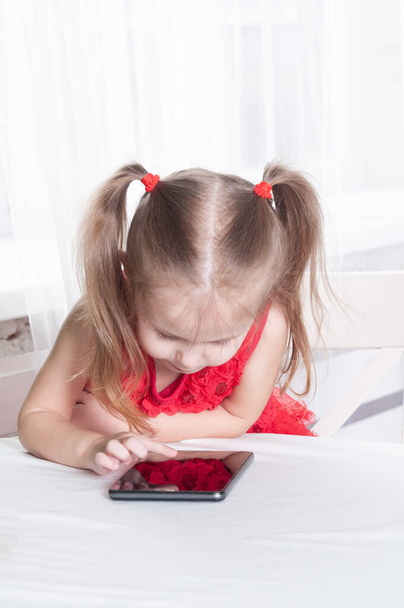 little girl in a red dress sits at a table and plays with a tablet, with a smile on her face - Fotoğraf, Görsel