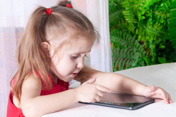 little girl in a red dress sits at a table and plays with a tablet, with a smile on her face - Foto, Imagem