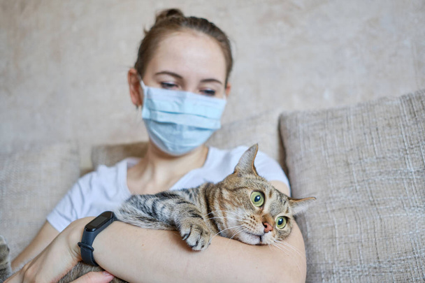 Das Mädchen mit der medizinischen Maske ist zu Hause auf sich allein gestellt und kümmert sich um die Katze. Nahaufnahme. - Foto, Bild