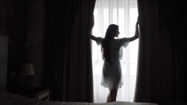 Hochzeitsmorgen der Braut. Ein schönes sinnliches europäisches Mädchen öffnet den Vorhang und genießt das Sonnenlicht, während es auf den Bräutigam wartet. Raffinierte natürliche Schönheit und Weiblichkeit. Silhouette einer Frau - Filmmaterial, Video