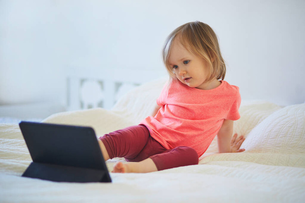 Toddler girl with digital tablet at home. Child watching cartoons. Kid using gadget to communicate with friends or kindergartners. Education and distance learning for kids. Stay at home entertainment - 写真・画像