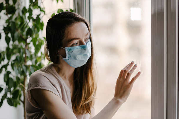 Steril tıbbi maskeli bir kız pencereden dışarı bakar ve koronavirüs salgını sırasında evde karantina ve kendini izole eder. - Fotoğraf, Görsel