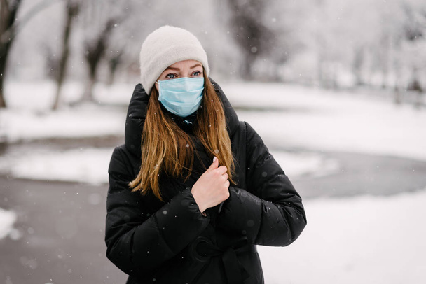 Девушка в стерильной медицинской маске от вирусов прячется от холода, завернувшись в черное пальто. Зимний день. На улице снег.
 - Фото, изображение