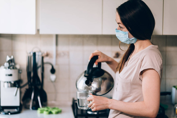 Μια γυναίκα με μάσκα κατά του ιού της στέψης στην κουζίνα ετοιμάζει τσάι για τον εαυτό της από κρυολογήματα και γρίπη για να μειώσει τη θερμοκρασία της. Κίνδυνος μόλυνσης από τον ιό - Φωτογραφία, εικόνα