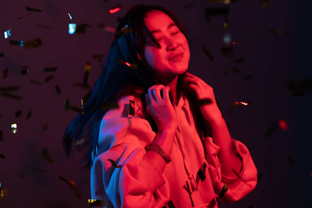 Excited asian attractive woman dancing, having fun, rejoices over confetti rain with neon light in studio. Concept of happiness, party, winning - Foto, immagini