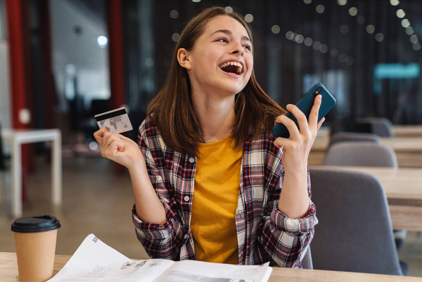 Image of delighted cute girl holding credit card and cellphone while doing homework in college library - Foto, imagen