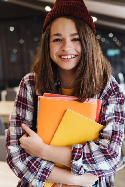 Image of joyful caucasian girl in hat smiling while holding exercise books in college library - Photo, image