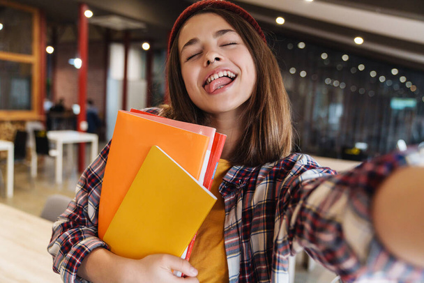 Image of amusing caucasian girl in hat taking selfie while holding exercise books in college library - Photo, Image