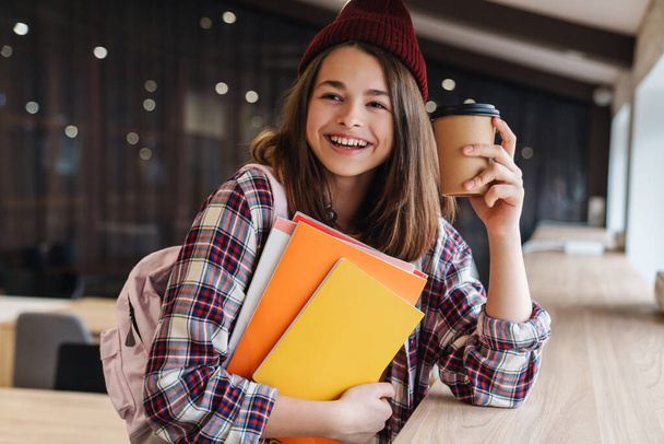 Image of joyful caucasian girl in hat drinking coffee while holding exercise books in college library - Фото, зображення