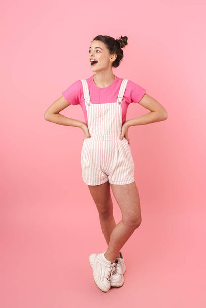 Photo of excited charming girl in overalls posing and expressing surprise isolated over pink background - Foto, Imagem
