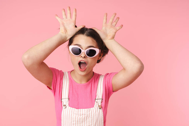 Photo of joyful charming girl in sunglasses making fun with hands isolated over pink background - Zdjęcie, obraz