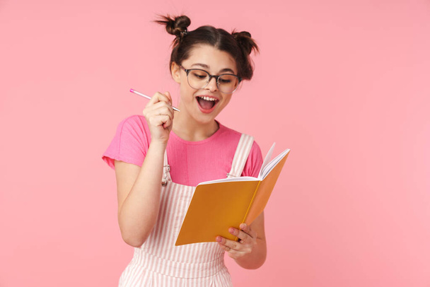 Photo of excited charming girl in eyeglasses writing in exercise book isolated over pink background - Zdjęcie, obraz