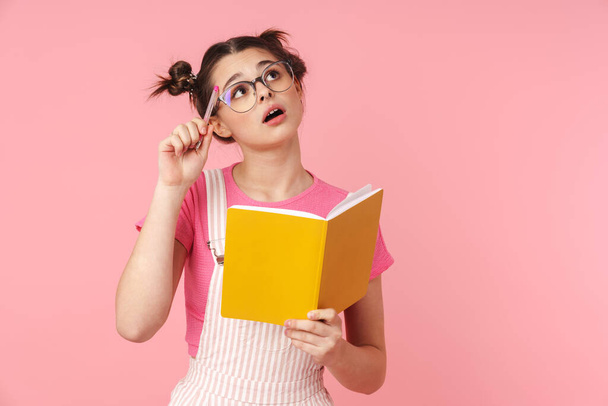 Photo of serious charming girl in eyeglasses holding exercise book and thinking isolated over pink background - Valokuva, kuva