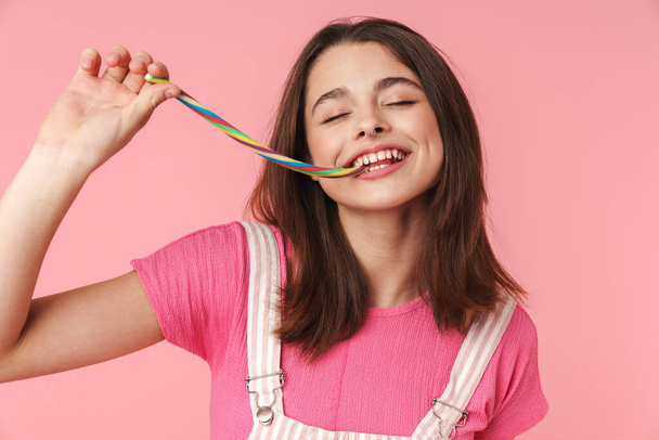 Photo of cheerful charming girl smiling and eating multicolored marmalade isolated over pink background - Foto, afbeelding