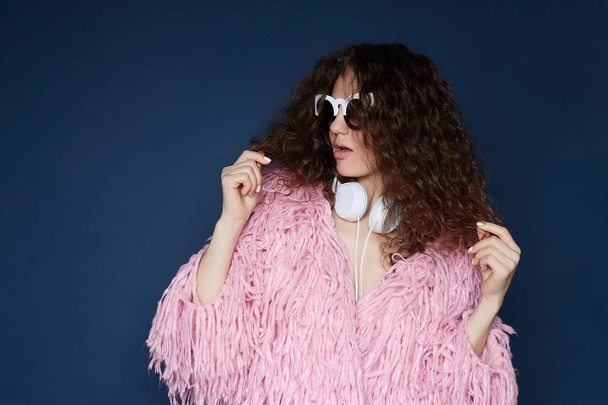 Close up shot of confident posh American female model wears trendy sunglasses, dressed in pink shaggy jacket, prepares for date, isolated on ultramarine wall, has fashionable look, listening to audio. - Photo, Image