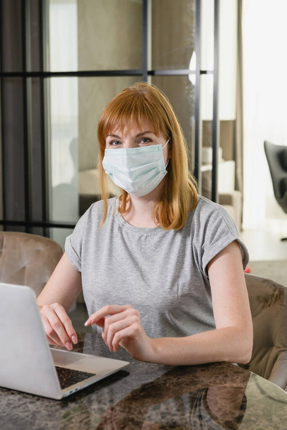 Woman sit on desk with open computer, using medical mask, during isolation period staying at home, study, work online. - Fotografie, Obrázek