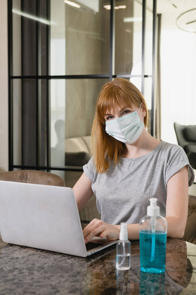 Γυναίκα κάθονται στο γραφείο με ανοιχτό υπολογιστή, χρησιμοποιώντας ιατρική μάσκα, κατά τη διάρκεια της απομόνωσης διαμονή στο σπίτι, μελέτη, εργασία σε απευθείας σύνδεση. - Φωτογραφία, εικόνα