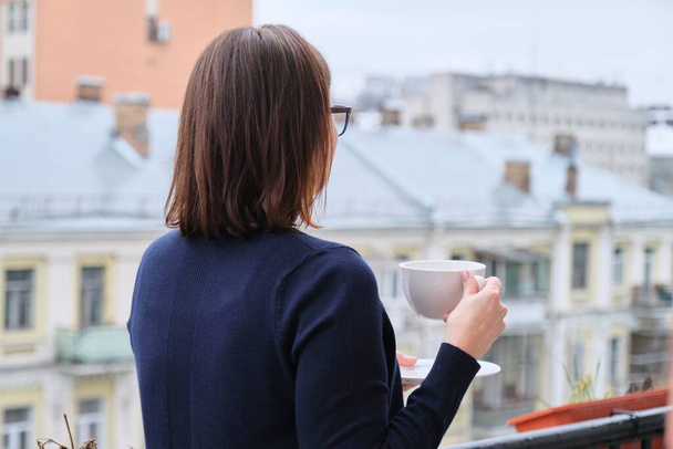 Mature woman standing on open balcony with cup of tea, female looks at city while in isolation, quarantined during viral infection, copy space - Foto, afbeelding