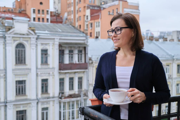 Portrait of mature woman in glasses, cardigan with cup of tea on open balcony in city. Background urban architecture, buildings, smiling female looks away, copy space - Fotografie, Obrázek