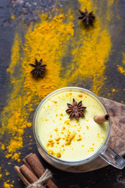 Golden milk or latte with turmeric (curcuma) powder with spices, dark brown concrete background. Trendy detox, immune boosting, anti-inflammatory healthy cozy drink.  - Foto, afbeelding
