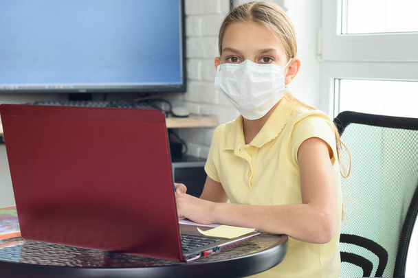 Ένα κορίτσι σε μια ιατρική μάσκα σε ένα σπιτικό περιβάλλον μαθαίνει εξ αποστάσεως και κοίταξε μέσα στο πλαίσιο - Φωτογραφία, εικόνα