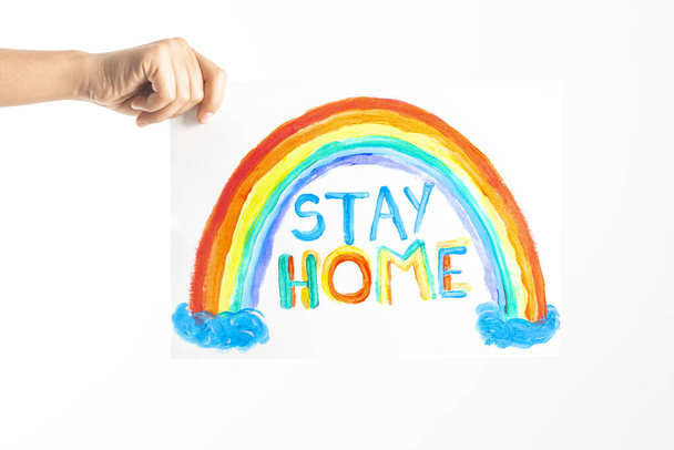 Mano de niño sosteniendo dibujo con colorido arco iris y palabras Quédese en casa. Campaña de medios sociales para la prevención del coronavirus
 - Foto, Imagen