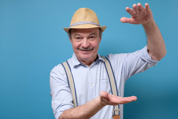 Senior ευτυχισμένος τρελός άνθρωπος σε καλοκαιρινό καπέλο δείχνει μεγάλο μέγεθος χειρονομία με τα χέρια, τονίζοντας μεγάλο ποσό από κάτι. Στιγμιότυπο στούντιο στον μπλε τοίχο - Φωτογραφία, εικόνα
