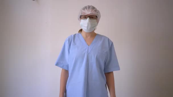 Female doctor in the protective mask and hat looks directly at the camera with white blank in hands. - Imágenes, Vídeo