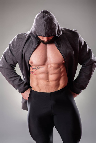 Seksikäs lihaksikas kehonbuider poseeraa huppari ja huppu harmaalla taustalla
 - Valokuva, kuva