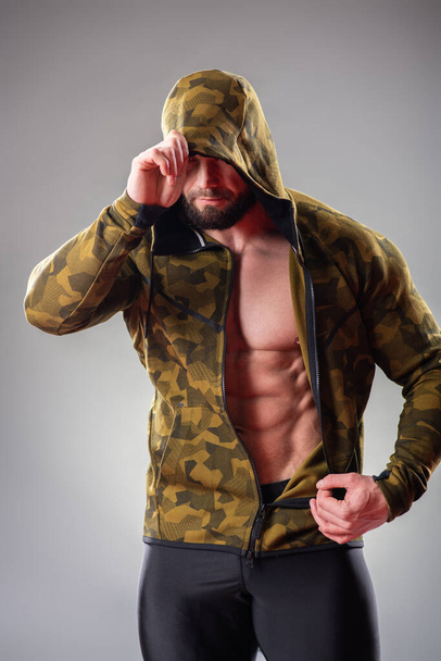 Sexy muscular bodybuider posing with sweatshirt and hood on the gray background - Foto, afbeelding