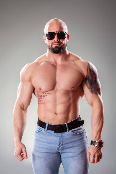 Sexy muscular bodybuider with sunglasses posing on the gray background - Fotó, kép