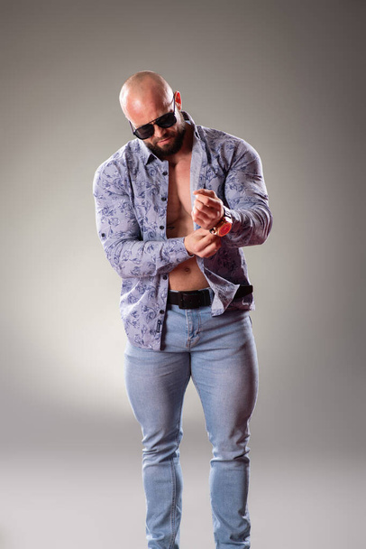 Sexy muscular bodybuider with sunglasses clothe his shirt on the gray background - Foto, afbeelding