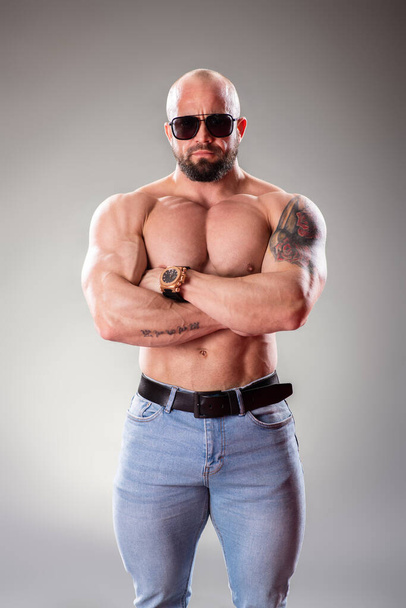Sexy muscular bodybuider with sunglasses posing on the gray background - Foto, afbeelding