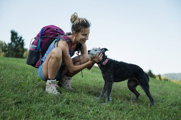 彼らは一緒にハイキングに行くとして彼女の黒い犬をパットする緑の牧草地にひざまずいてバックパックを持つ幸せな若い女性. - 写真・画像